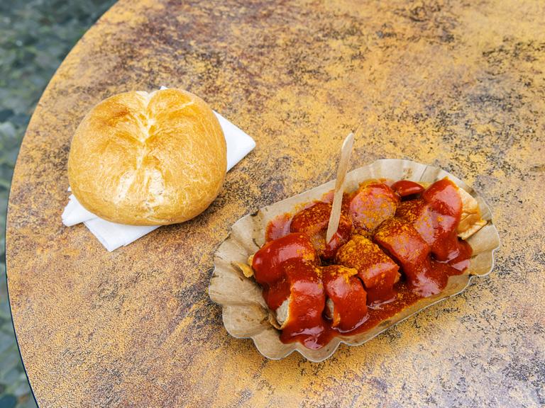 EIne Currywurst und ein Brötchen liegen auf einem Tisch vor Konnopkes Imbiss, dem Kult-Imbissstand im Berliner Stadtteil Prenzlauer Berg. Konnopke gilt als erster Imbissstand in Ost-Berlin, bei dem im Jahr 1960 die Currywurst eingeführt wurde.