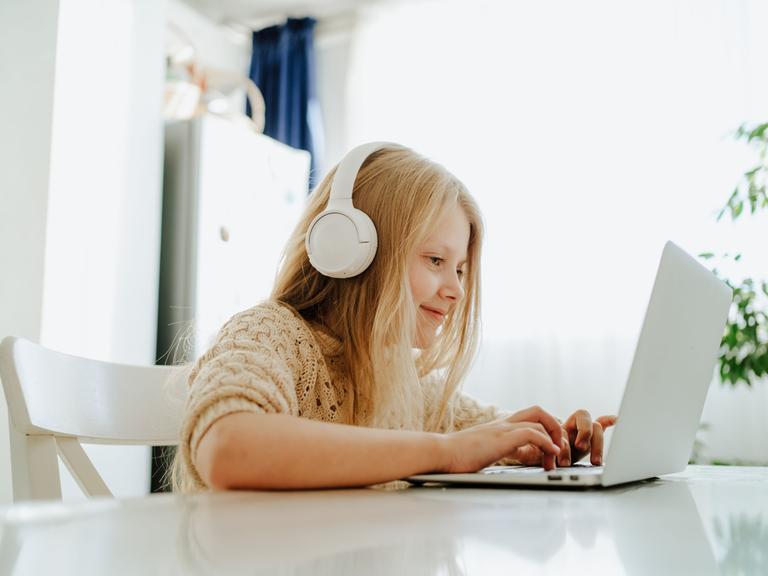 Ein Mädchen mit weißen Kopfhörern sitzt am Tisch vor einem Laptop und schreibt.